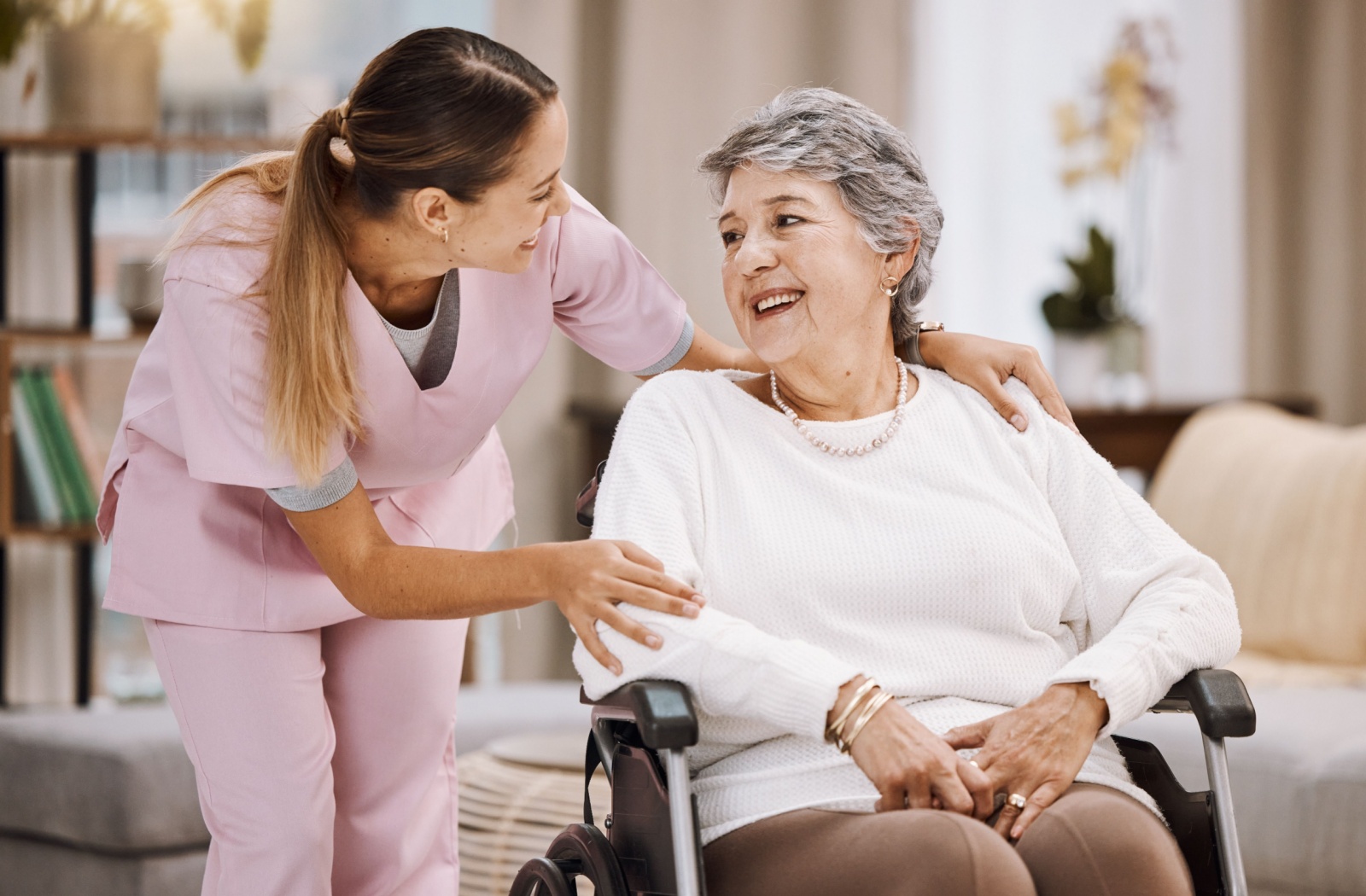 A caregiver rests her hands on the shoulders of a senior woman in a wheelchair as she checks in with her.