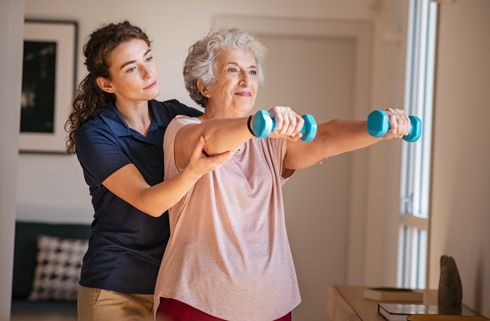 A physical therapist assists a senior in living 5-pound dumbbells straight up.