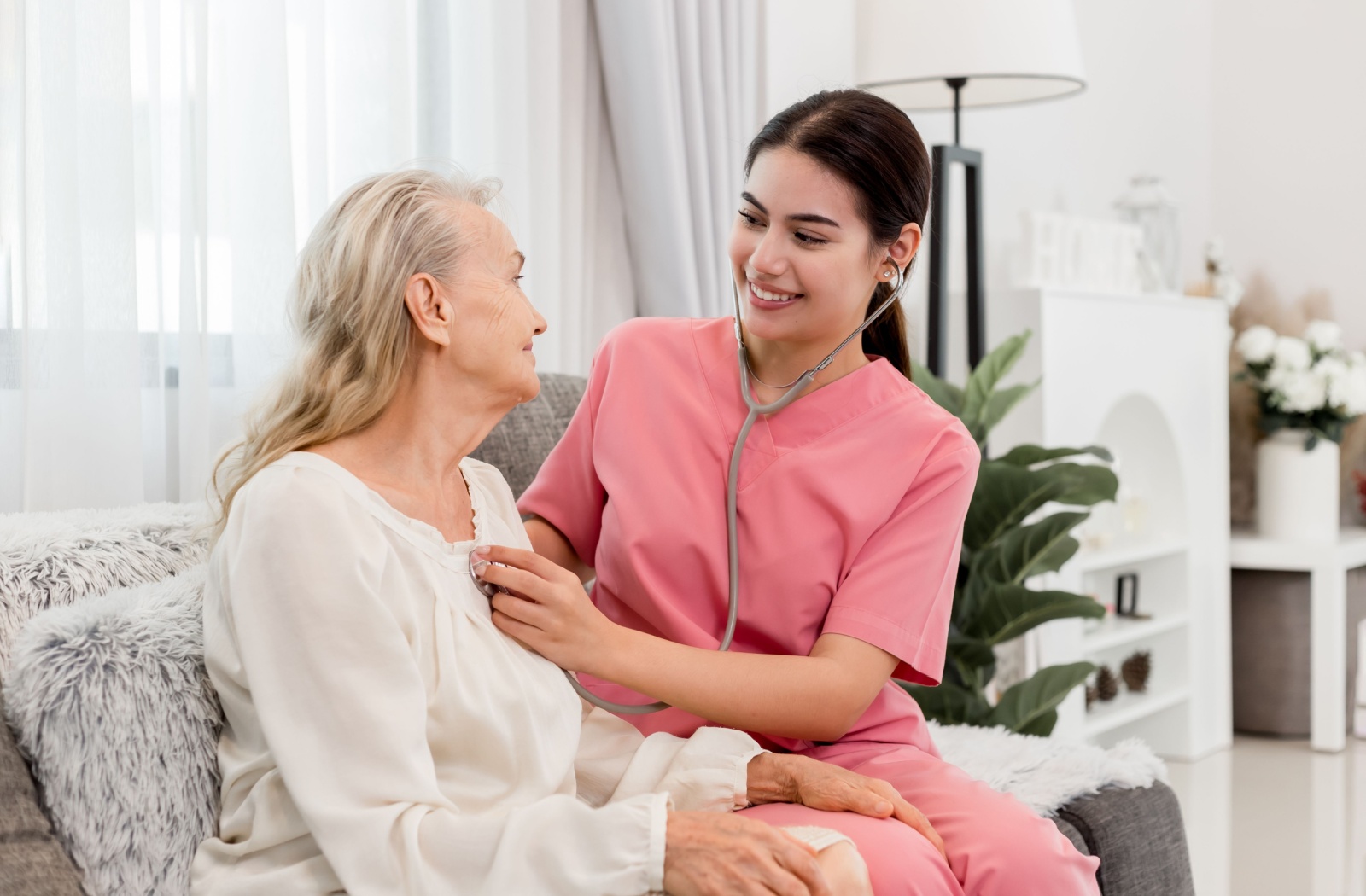 A smiling skilled nurse with a stethoscope sitting beside a senior resident on the couch and listening to her heart.