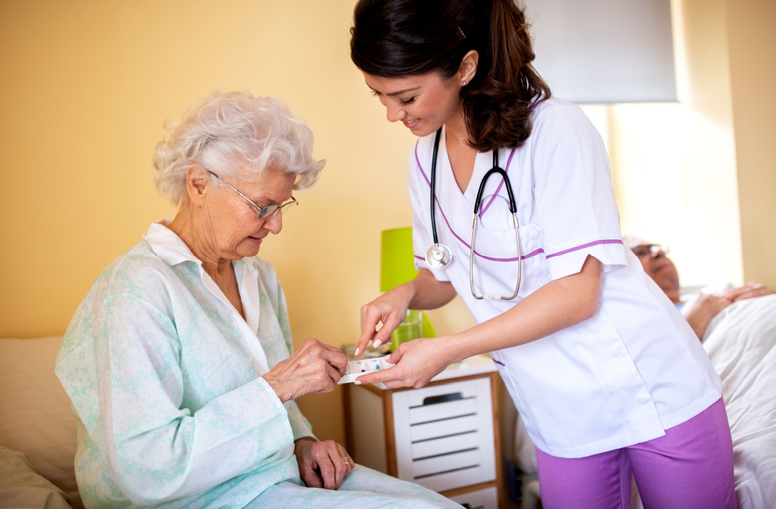 A smiling skilled nurse standing next to a senior resident sitting on a bed and handing her medication from a pill tray.
