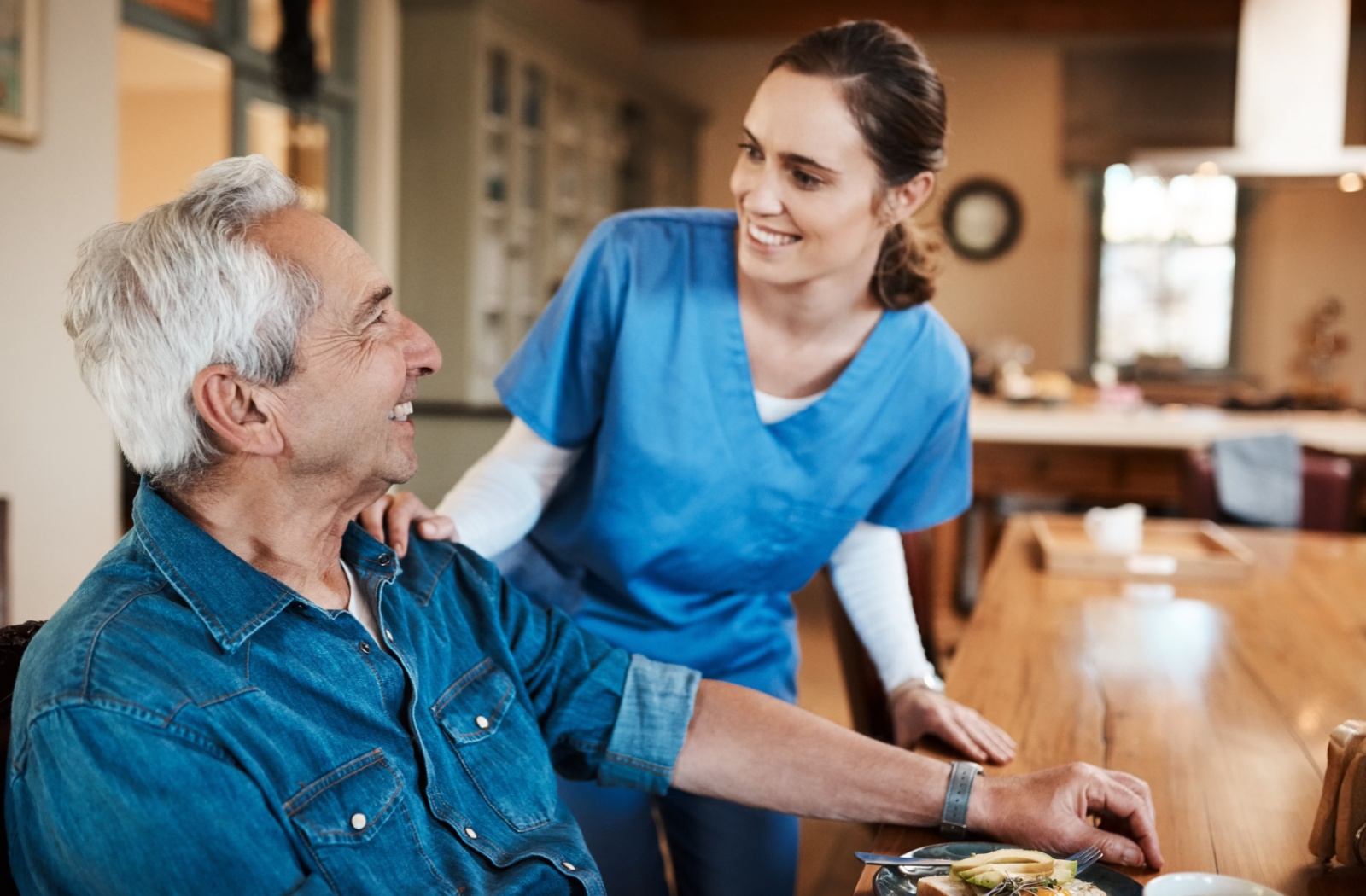 Older man receiving compassionate care from a respite caregiver, smiling and engaging in conversation at the dining table.