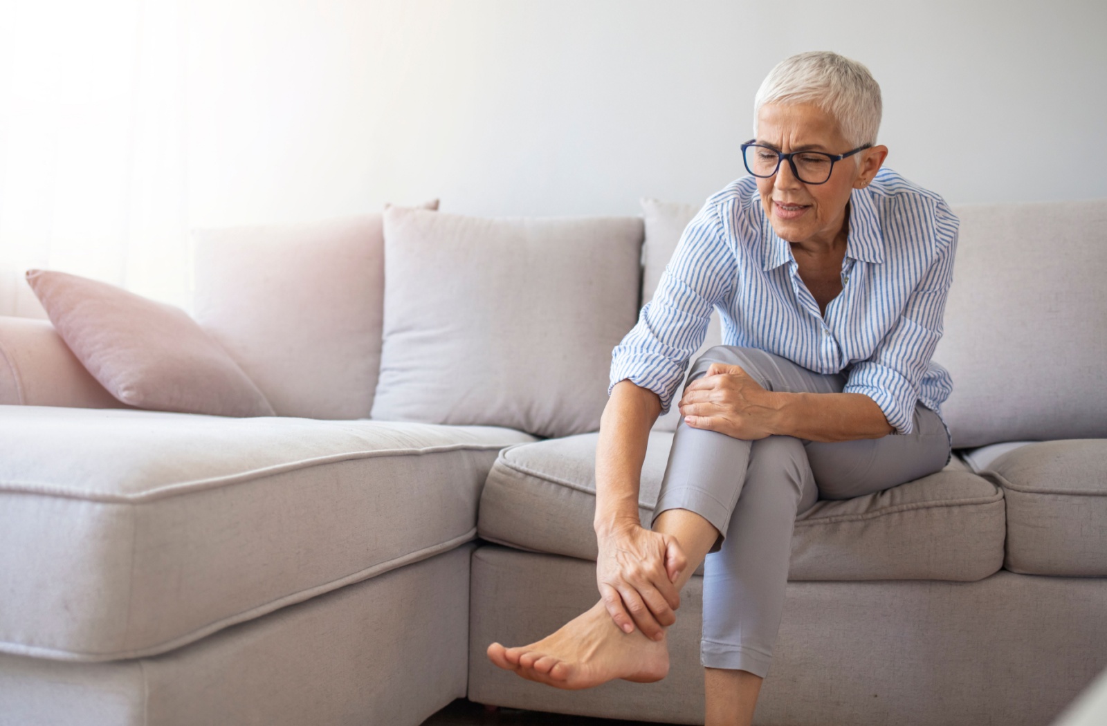 An older adult holds their ankle and knee while sitting on the couch to reduce pressure on their swollen ankle