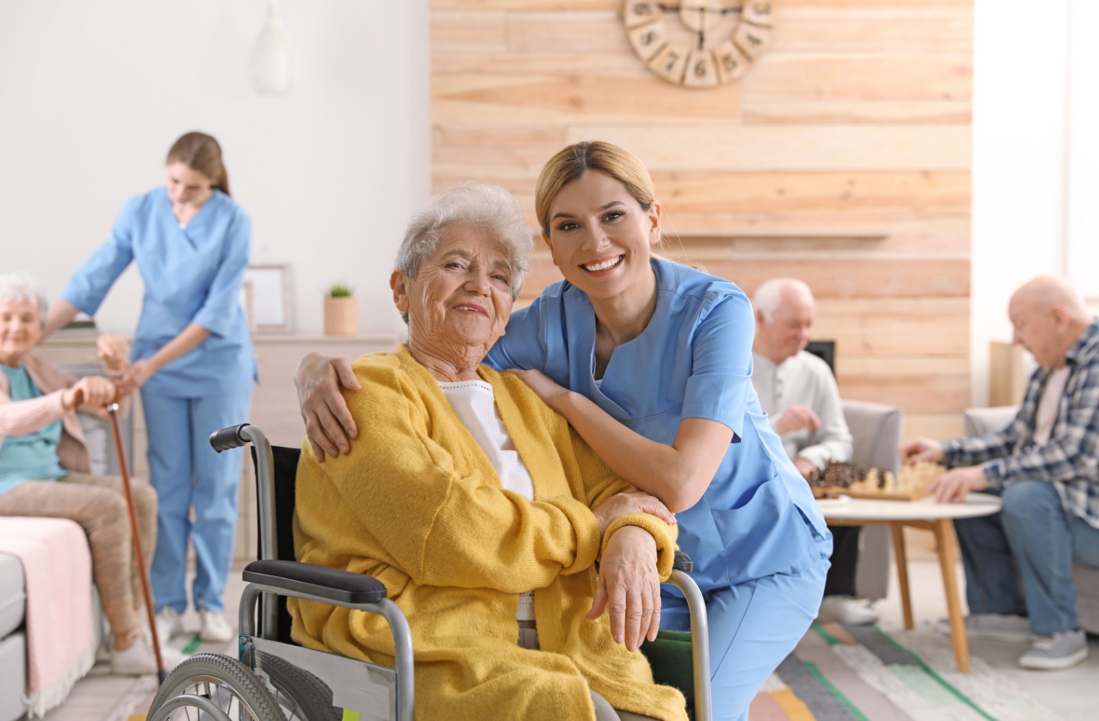 A caregiver kneels beside a resident in skilled nursing while other older adults play games in the background