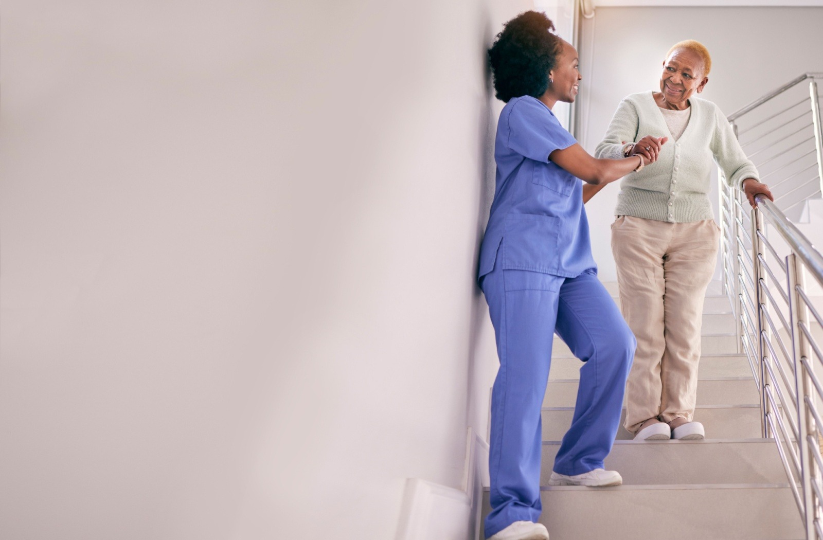 A caregiver assists a senior resident walking down a flight of stairs.
