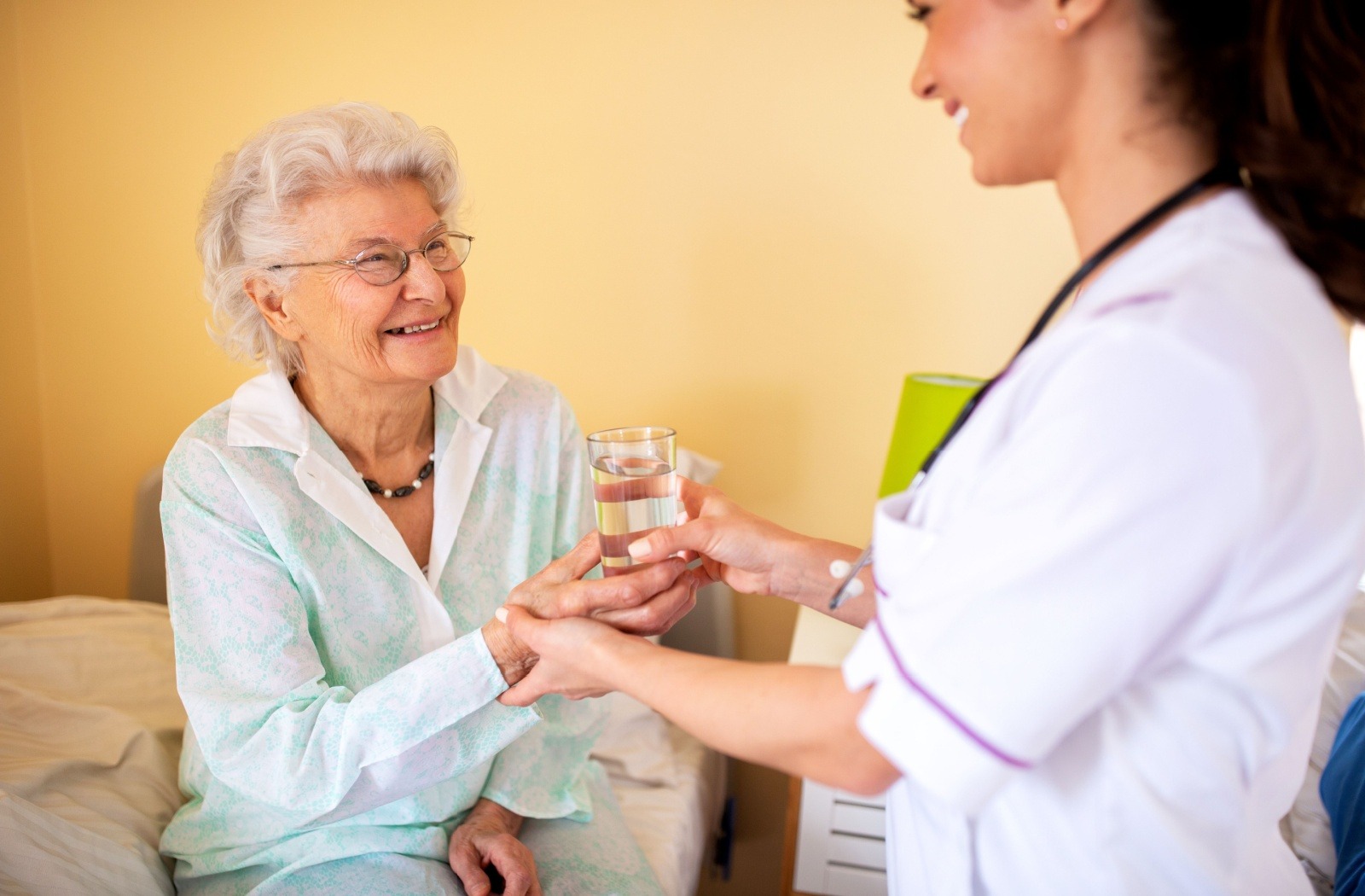 A nurse at a skilled nursing community hands a glass of water to a smiling senior sitting on a bed.
